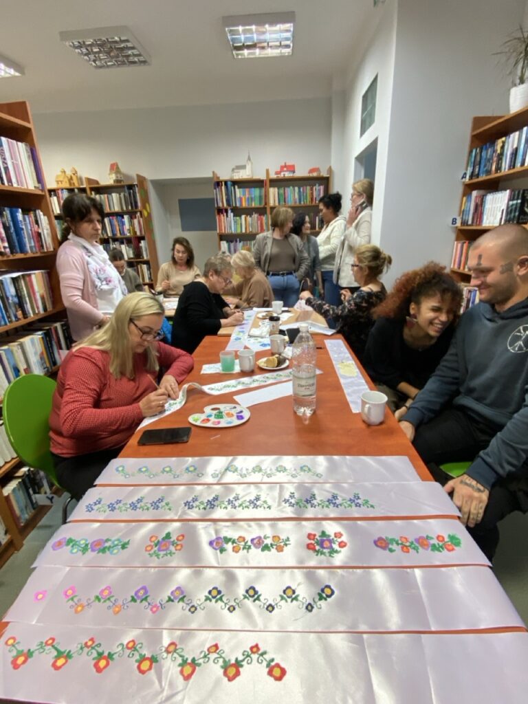 Pomalowane wstążki na stole a za nimi grupa osób. Jedni malują, inni rozmawiają.