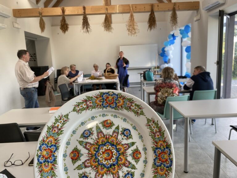 Talerz pomalowany w drobne ornamenty kolorowych kwiatków, a w tle grupa ludzi biorących udział w warsztatach