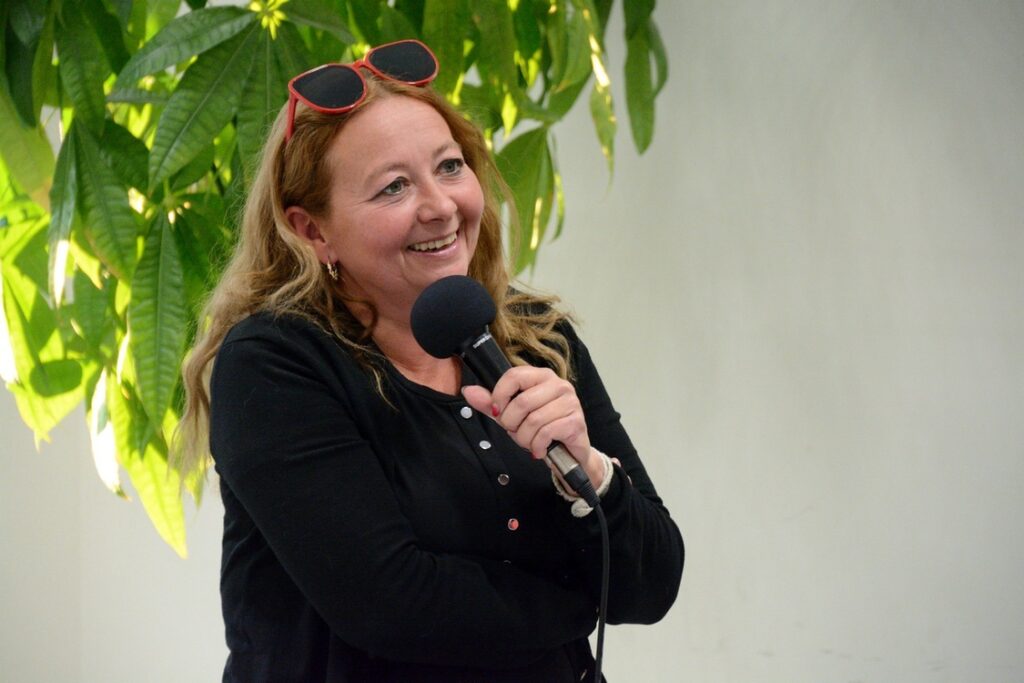 uśmiechnięta konbieta z jasnymi, falowanymi włosami stoi na tle ściany i trzyma w dłoni mikrofon