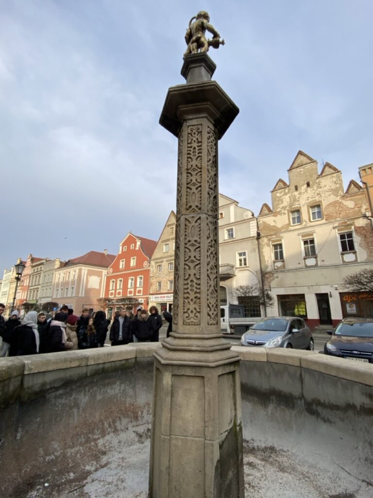 Fontanna zwieńczona rzeźbą złotego amorka, na jej tle grupa młodzieży.