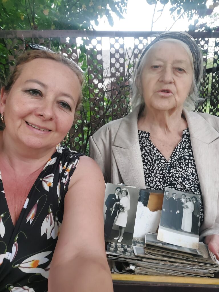 Dwie koniety. jedna młodsza, druga w zaawansowanym wieku. Siedzą przy stole przy starych zdjęciach