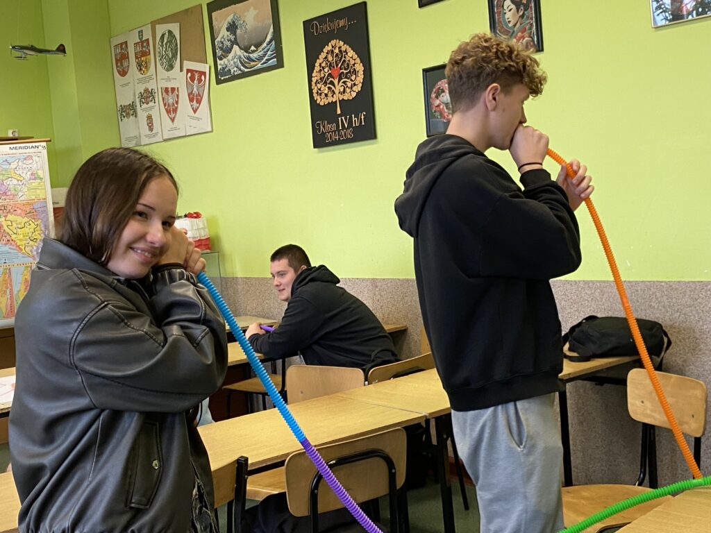 dziewczyna trzy kolorową rurkę przy uchu, uśmiecha się, obok niecj chłopak trzyma drugi koniec tej długiej rurki przy ustach. Dziewczyna uśmiecha się, a chłopak stoi do dziewczyny obrócony plecami. 