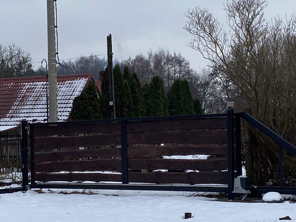 na zdjęciu jest brama wjazdowa w czarnej ramie osadzone poziome drewniane , ciemnobrązowe deski