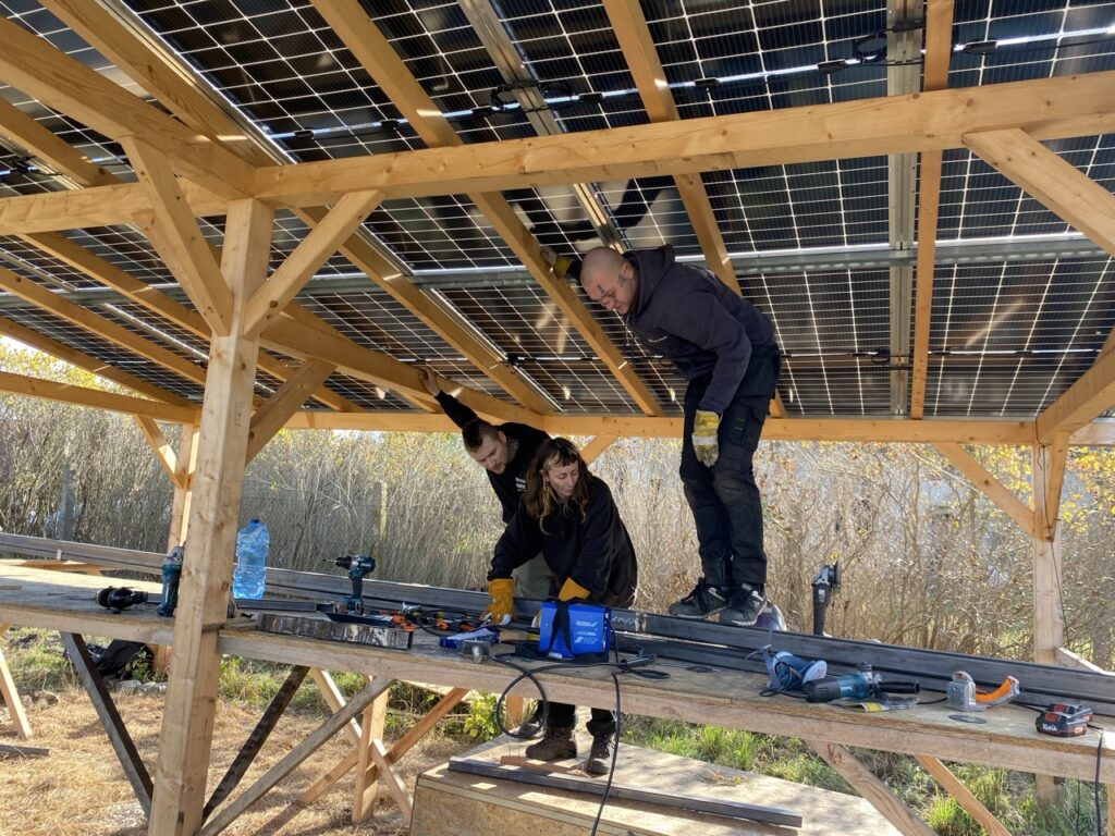 pod drewnianym dache z panelami fotowoltaicznymi stoją na dużym stole roboczym mężczyźni i kobieta przy pracy