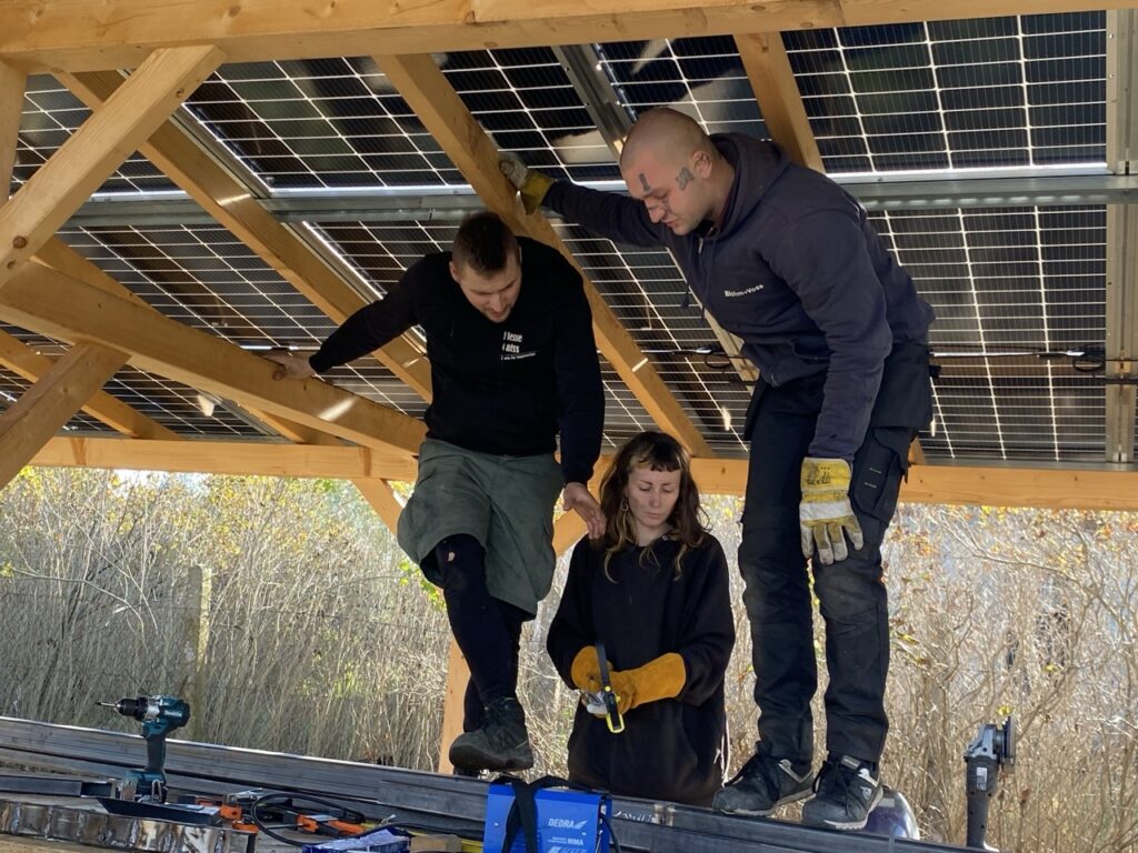 pod drewnianym dache z panelami fotowoltaicznymi stoją na dużym stole roboczym mężczyźni i kobieta przy pracy