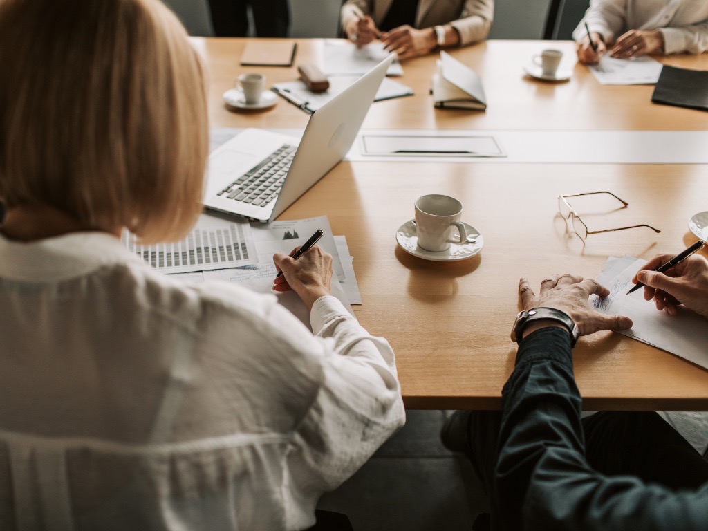 stół, na stole stoi laptop, filiżanki z kawą, dokumenty, przy stole siedzi tyłem kobieta oraz grono osób, których dłonie widac na stole