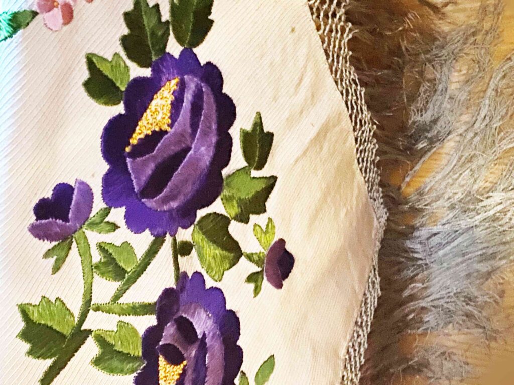 kawałek chusty spływający w dół, z długimi frędzlami wyszyty w fioletowe róże na zielonej gałązce z płatkami