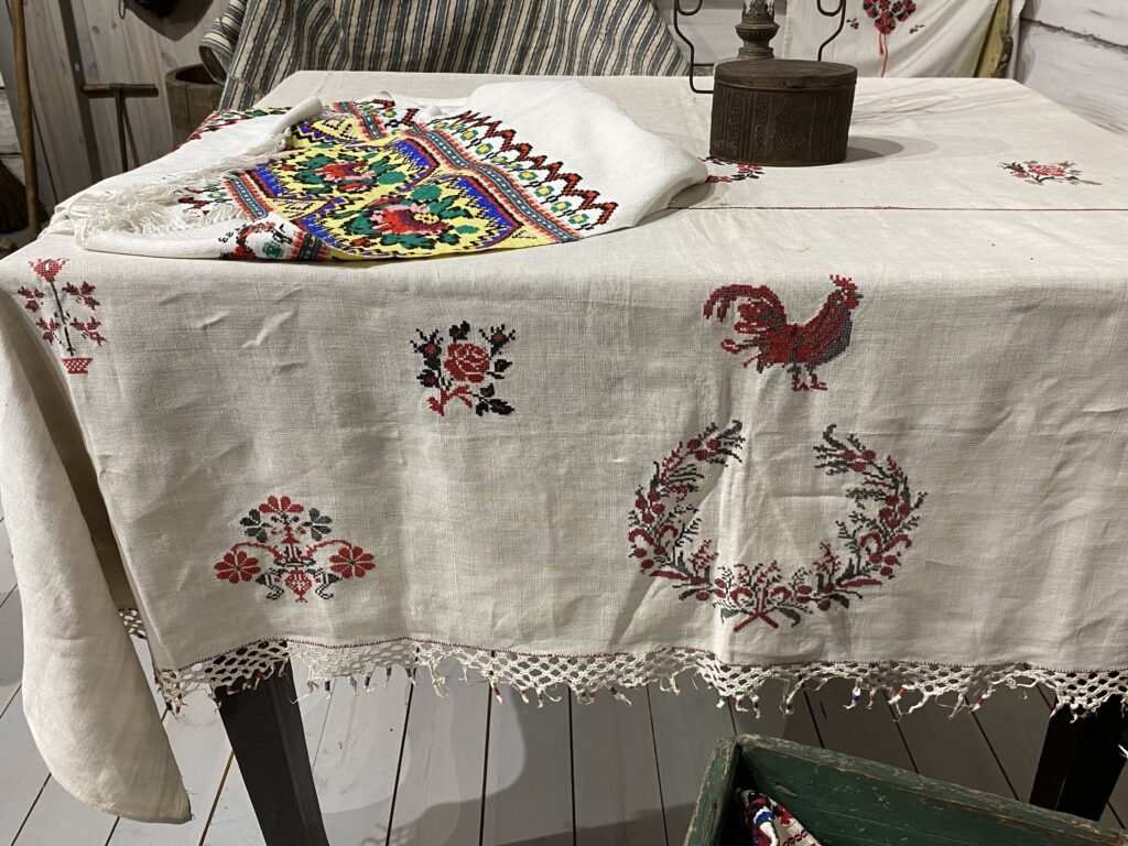 stół nakryty obrusem z naturalnego koloru wyszytym w motywy krzyzykowe w kolorach czerwono-czarnym, na stole leż bieżnik w innym, kolorowym haftem krzyżykowym - geometrycznym.