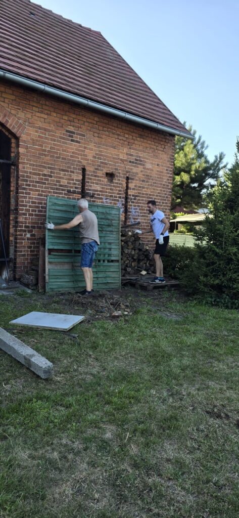 Dwóch mieszkańców przenosi drewniane elementy podczas porządkowania terenu wokół budynku przekazanego lokalnej społeczności w Lędzinach.