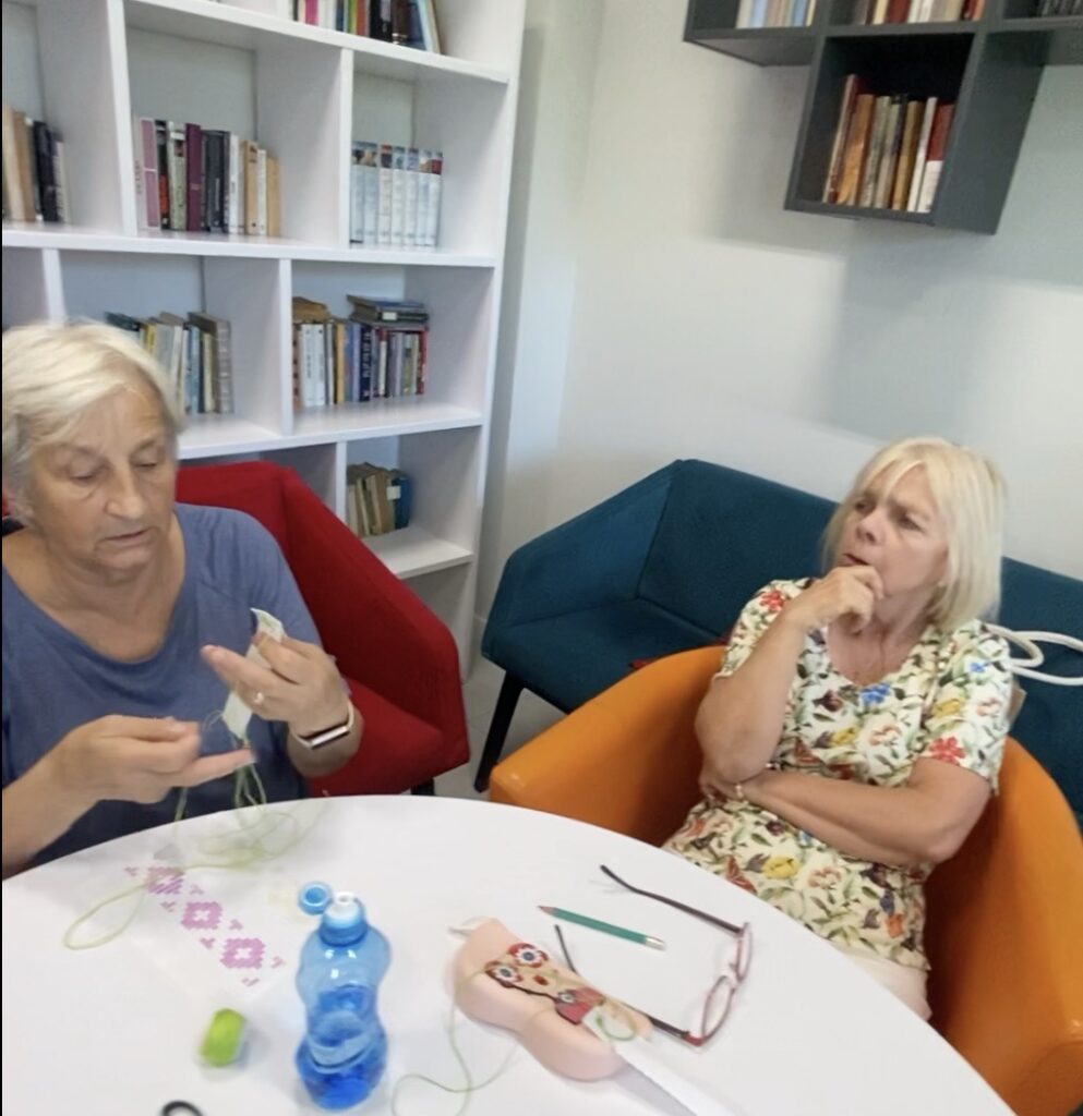 kobiety siedzą przy stolikach i wyszywają na pasku kanwy